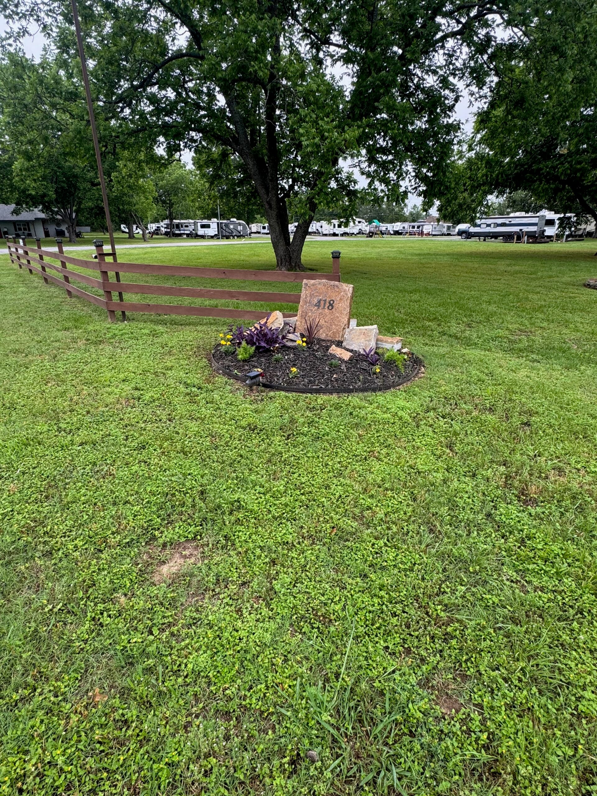 A landscaped area with decorative stones, one marked "418," sits near a wooden fence on a grassy lawn with trees and RVs in the background.