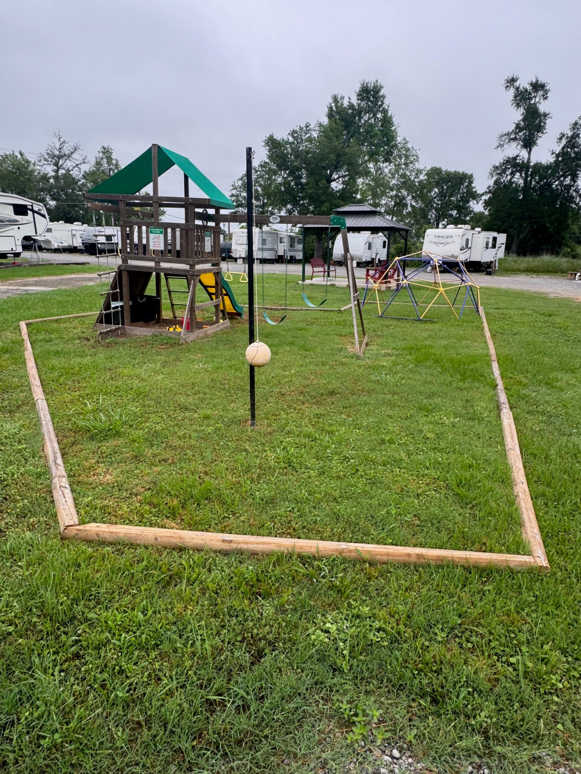 A playground area with a wooden playset, swings, geometric climbing dome, and a tetherball pole, surrounded by grass and bordered by wooden logs, with RVs parked in the background.