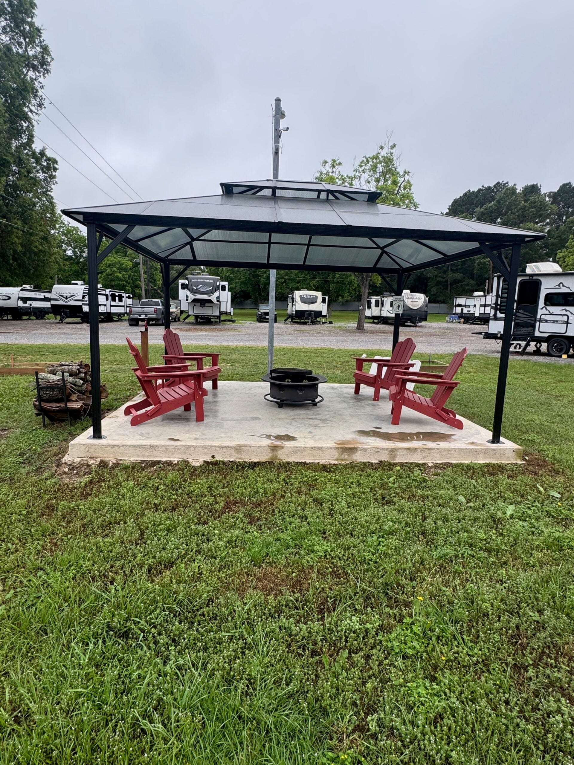 A covered gazebo with four red Adirondack chairs arranged around a fire pit on a concrete pad, surrounded by grass and RVs in the background.