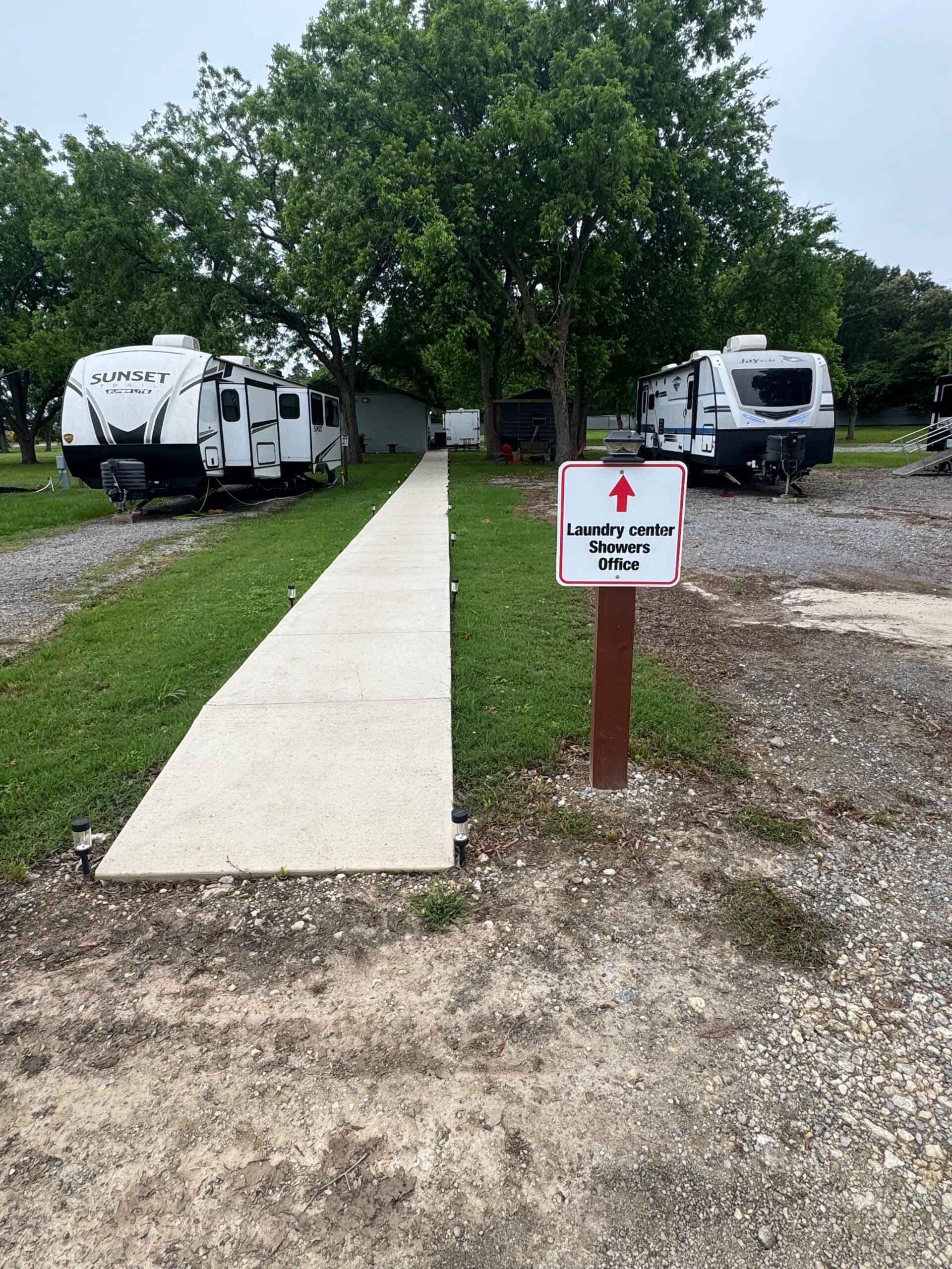 A concrete walkway leads between two RVs toward a building, with a sign indicating the direction to the laundry center, showers, and office.