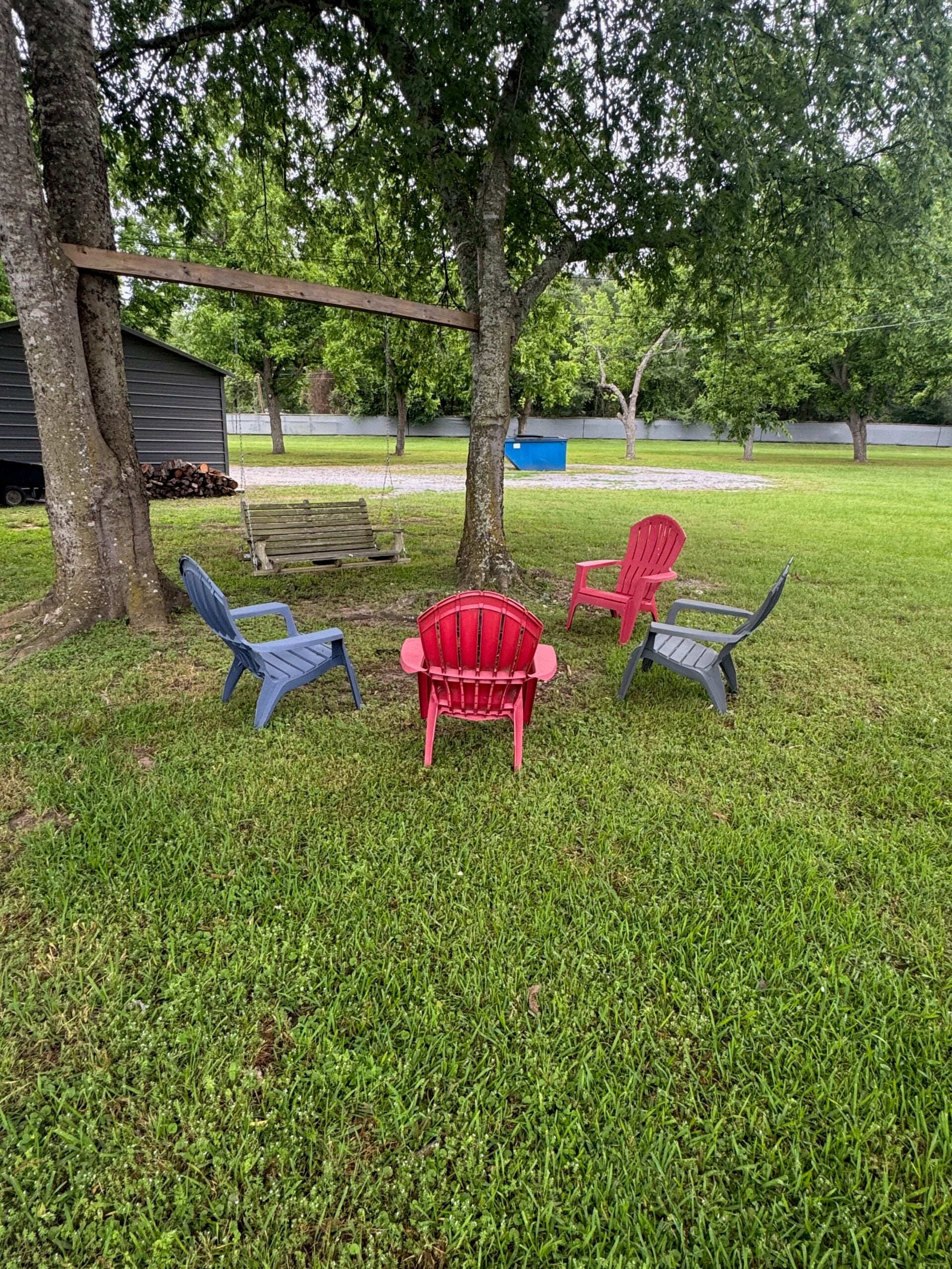 Four Adirondack chairs, three gray and one red, are arranged in a circle on a grassy lawn near a wooden swing and trees in a backyard setting.