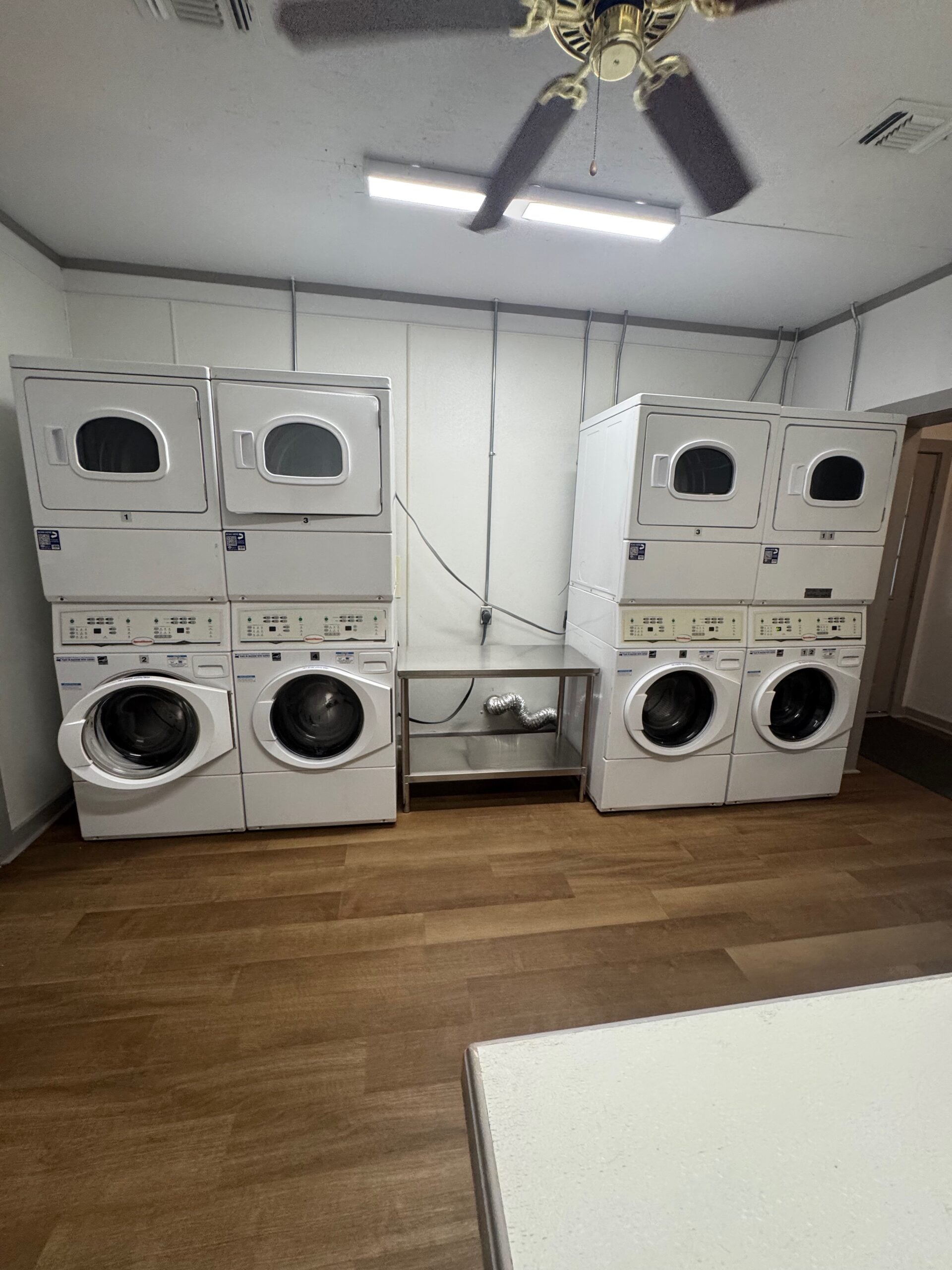 A laundry room with four stacked washer and dryer units against the wall, wood-look flooring, and a ceiling fan.