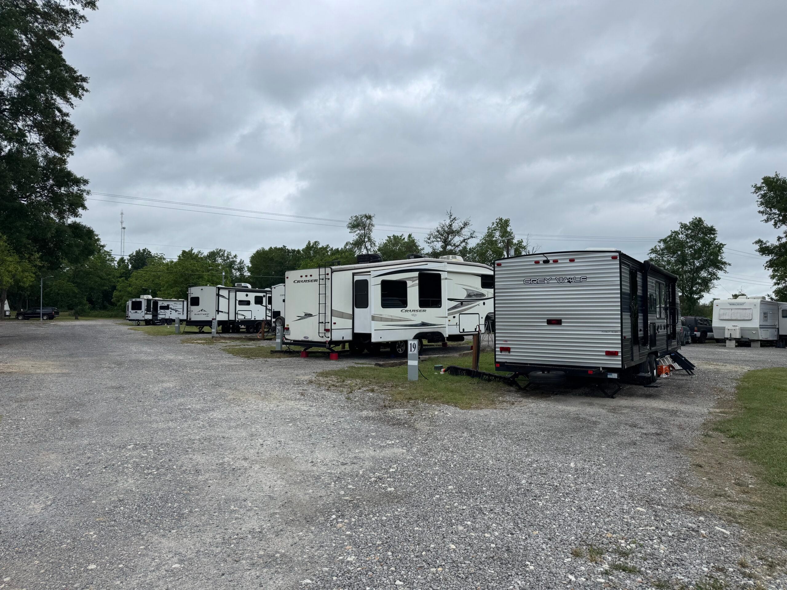 Several RVs are parked on gravel lots in a campground, surrounded by trees under a cloudy sky.