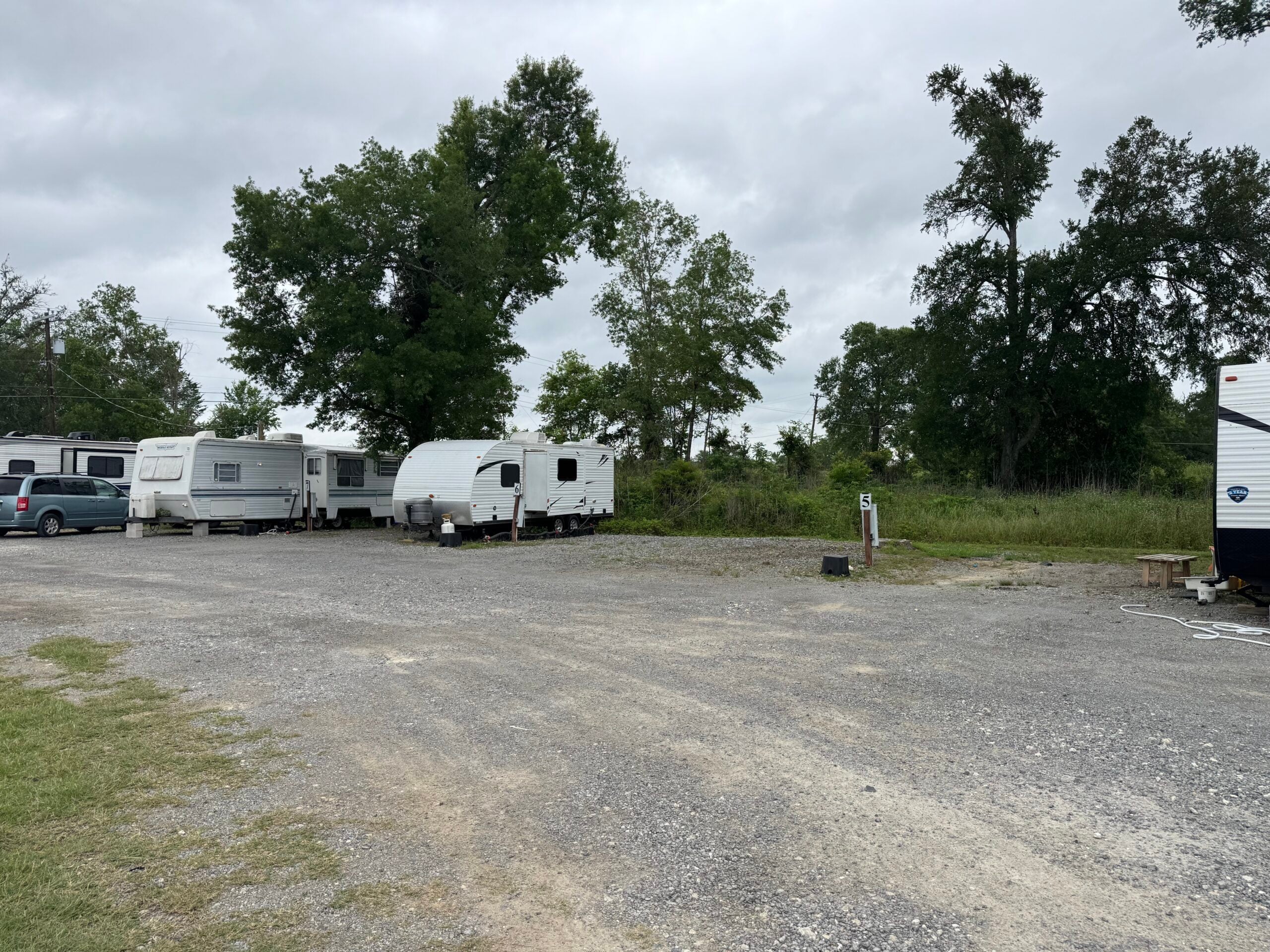 Several RVs are parked on a gravel lot bordered by grass and trees under a cloudy sky.