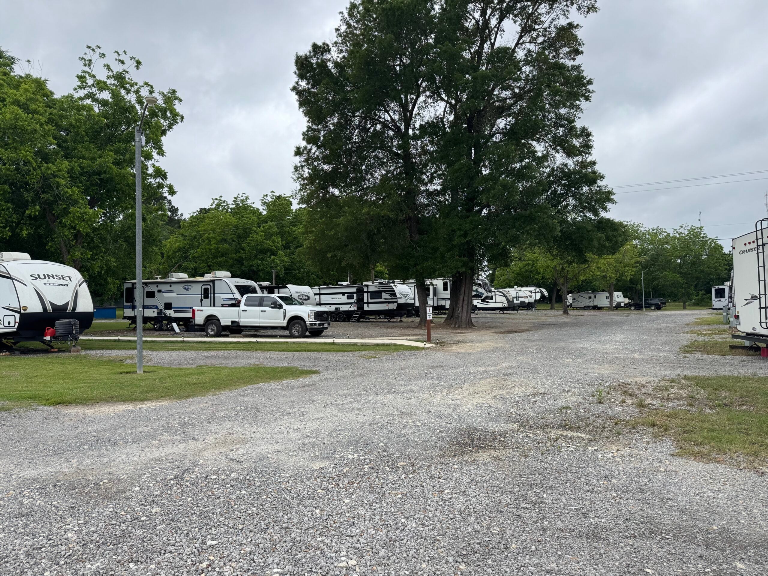 A gravel lot with several parked RVs and a white pickup truck, surrounded by green trees under a cloudy sky.